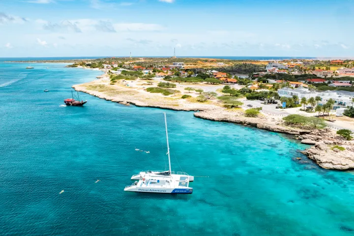 Catamaran in turquoise sea near rocky coast with buildings and greenery under blue sky.