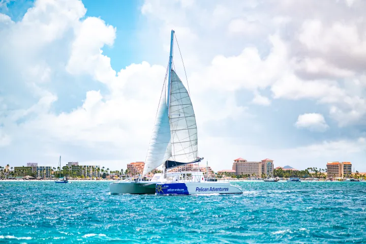 Catamaran sailing on turquoise sea with buildings in the background.