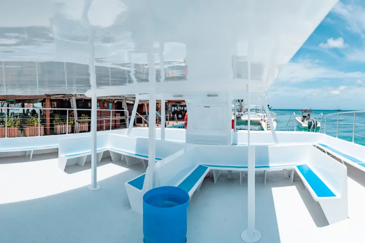 Empty ferry deck with white seats and blue accents, docked at a sunny, tropical waterfront.