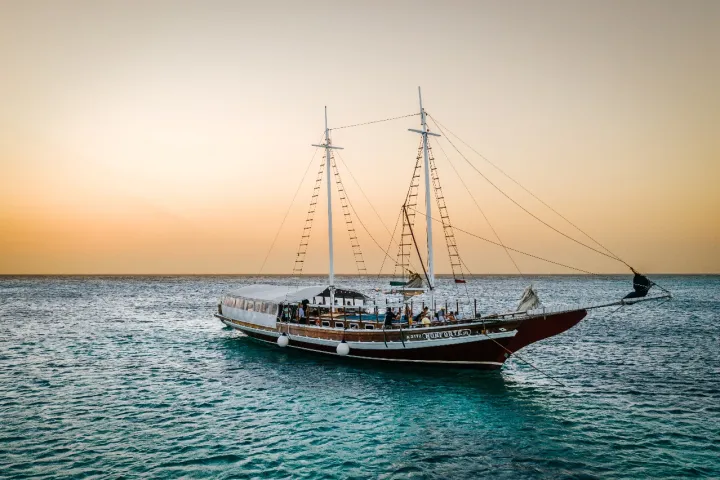 Sailing ship on calm ocean at sunset with orange sky.