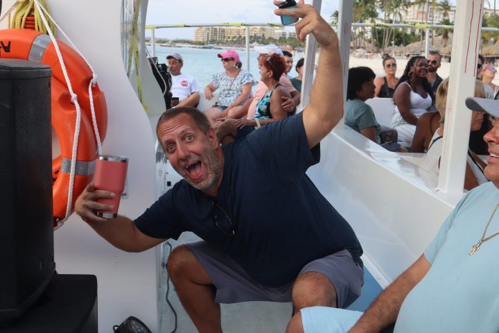 Man joyfully posing on a boat with a drink, surrounded by seated people.