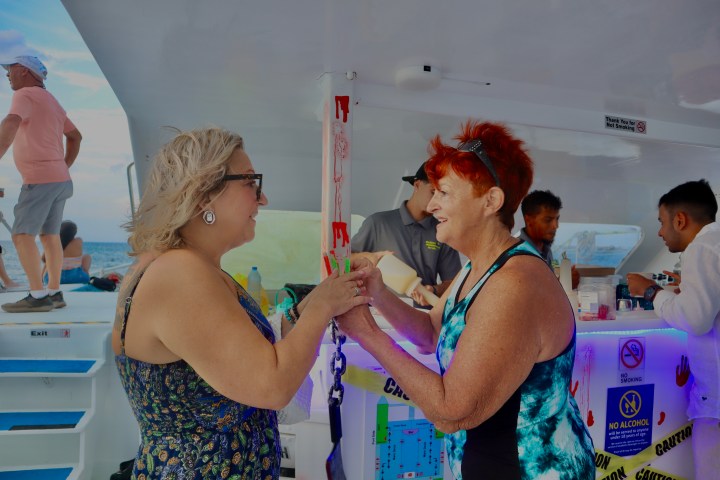 Two women chatting and smiling on a boat near a bar with people and ocean in the background.