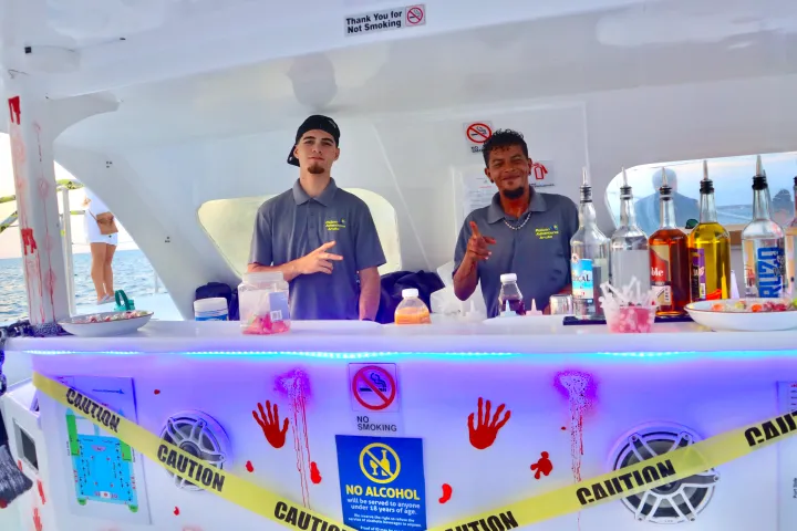 Two men behind a beachside bar with bottles and 'Caution' tape, decorated with handprints.