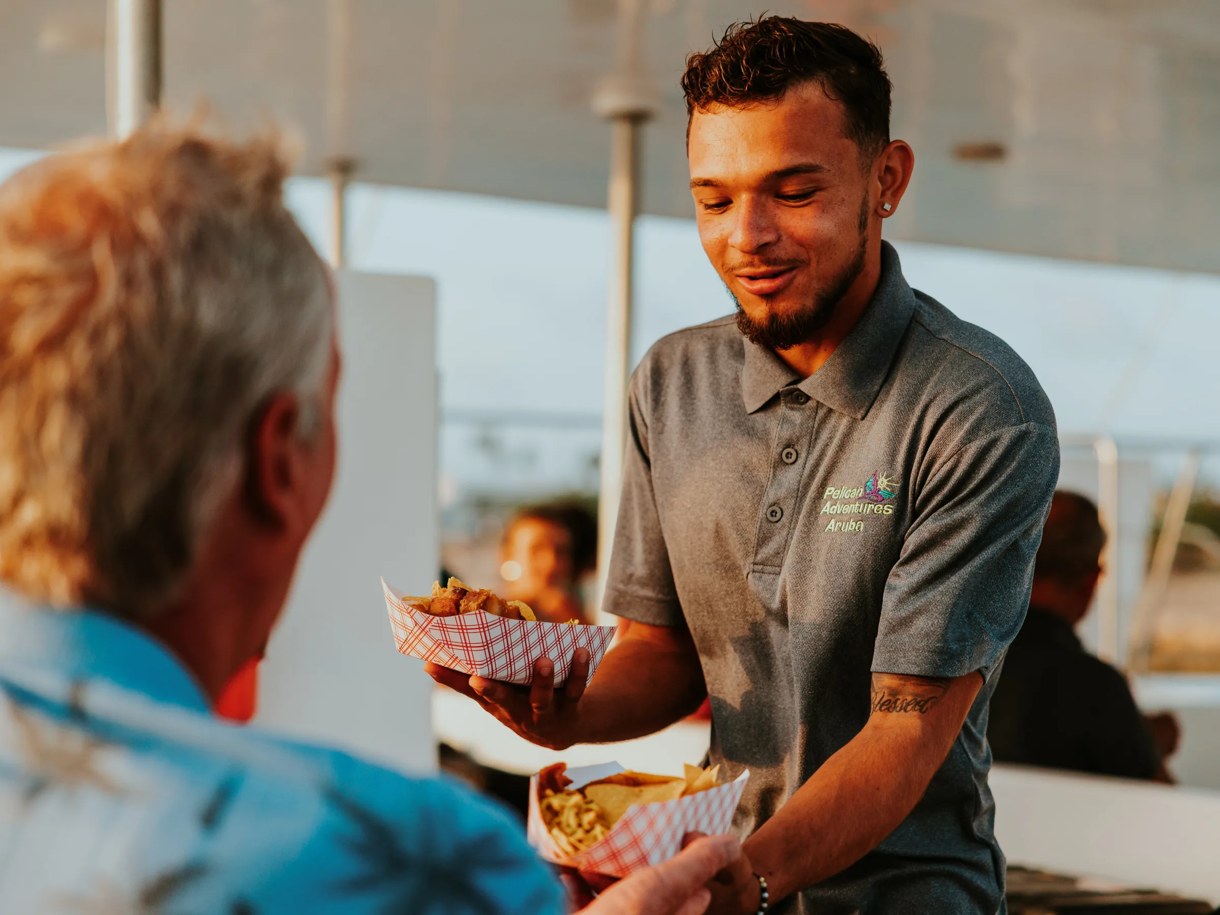 Smiling server handing food trays to a seated customer on a sunny day.
