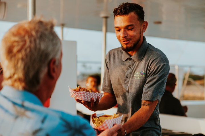 Smiling server handing food trays to a seated customer on a sunny day.