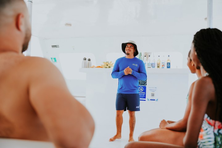 Guide in blue shirt speaking to seated people on a boat with drinks in the background.