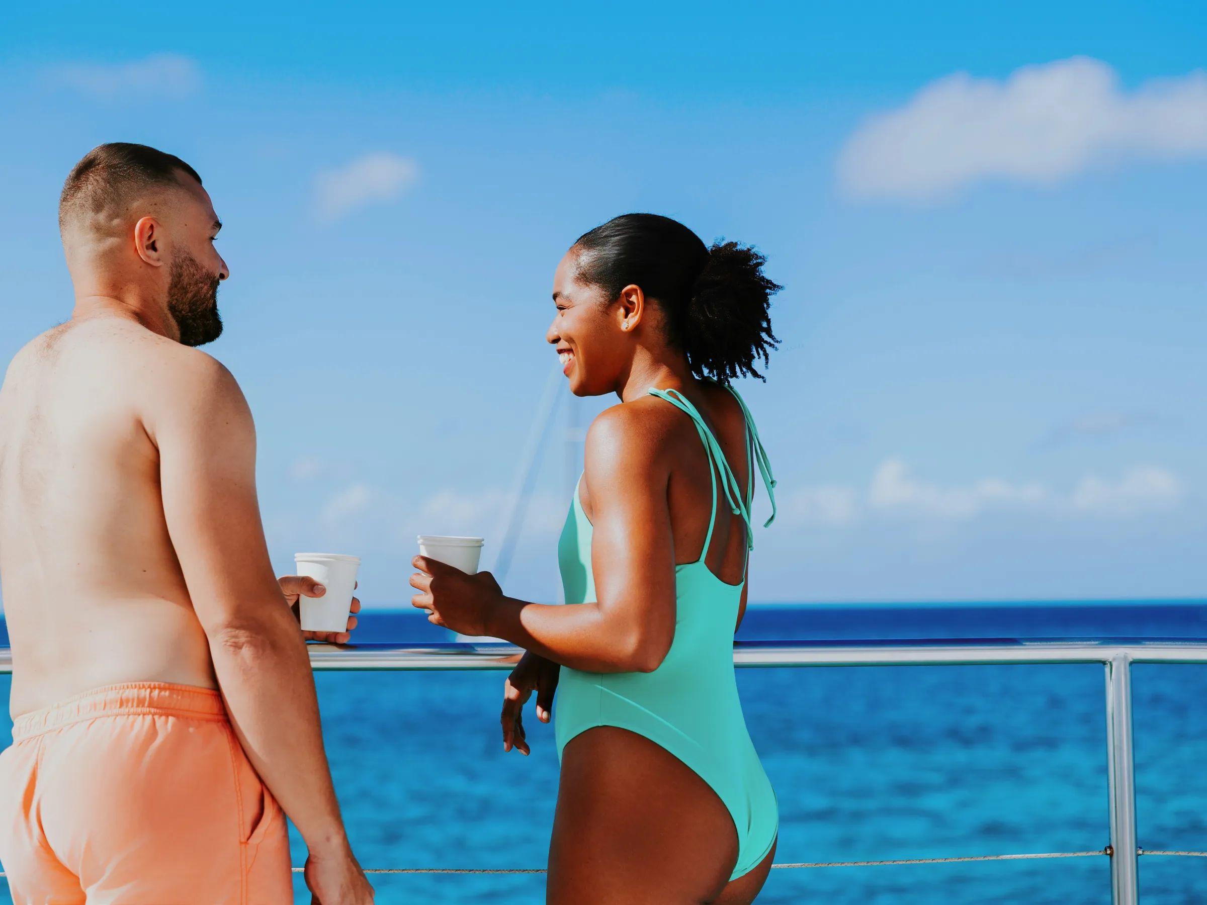 Two people on a boat deck with drinks, ocean view in background.