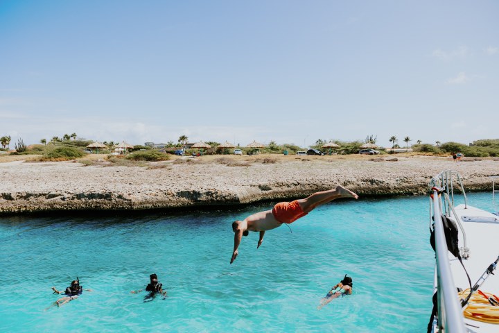 Person diving off boat into clear water, with snorkelers and rocky shore in background.