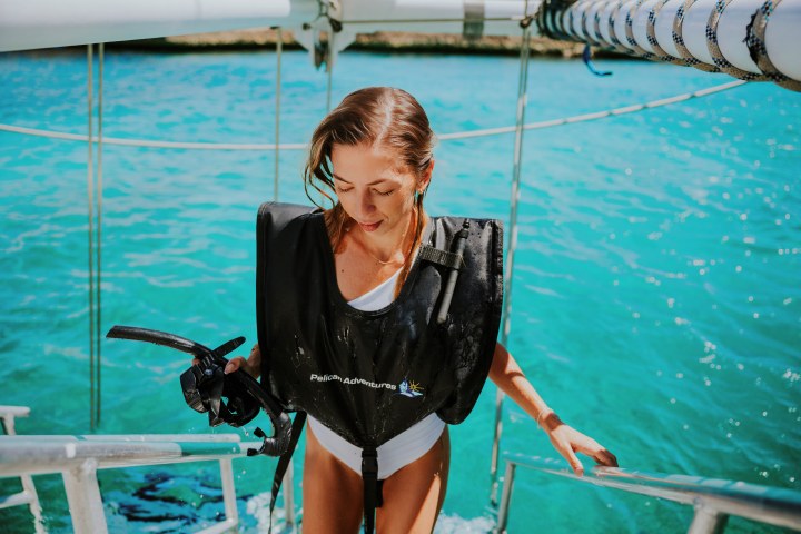 Woman in a life vest climbing boat ladder from clear blue water, holding snorkeling gear.
