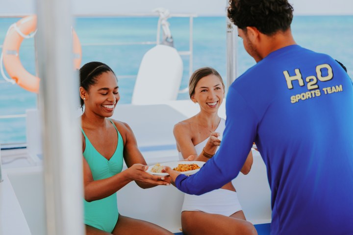 Two women on a boat smile as a man in a blue shirt serves them food.