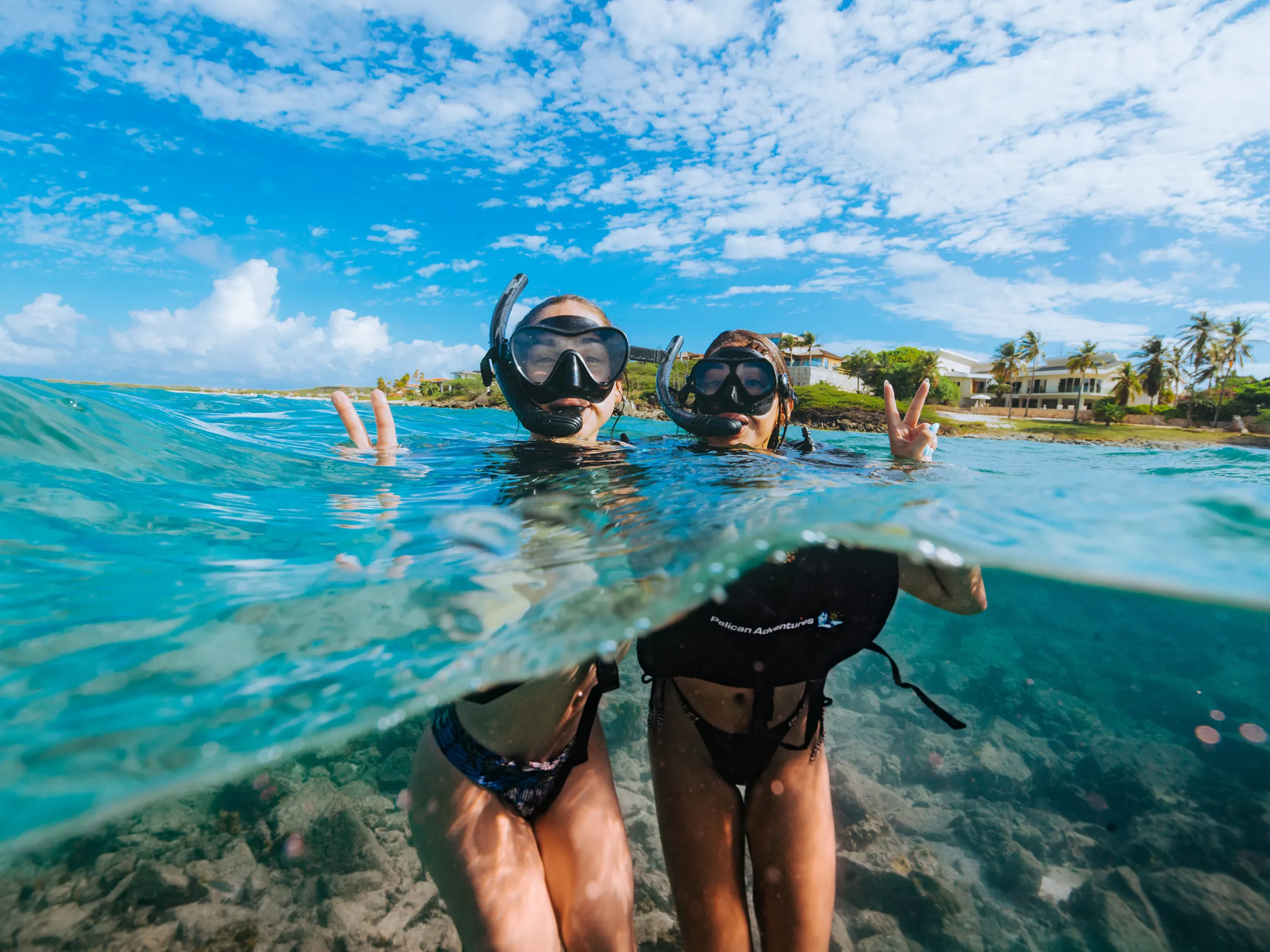 Two people snorkeling near the water surface, showing peace signs, with a clear sky and shoreline in the background.