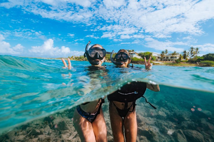 Two people snorkeling near the water surface, showing peace signs, with a clear sky and shoreline in the background.
