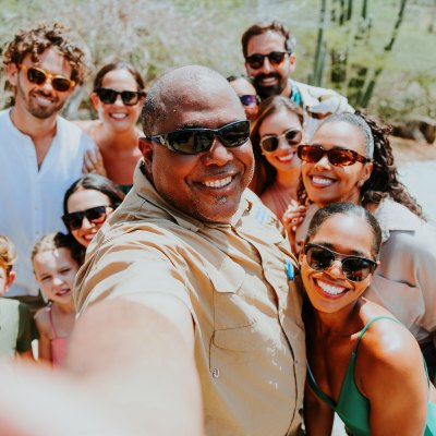 Group of nine smiling people taking a selfie outdoors, wearing sunglasses.