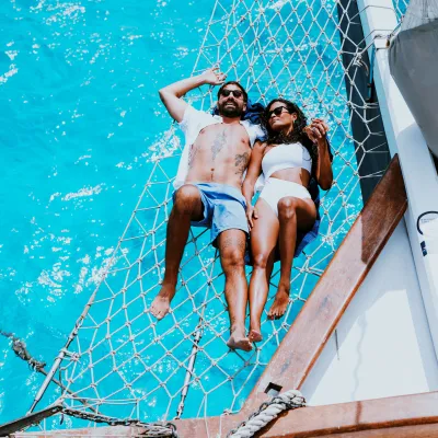 Two people relaxing on a boat net above turquoise water on a sunny day.