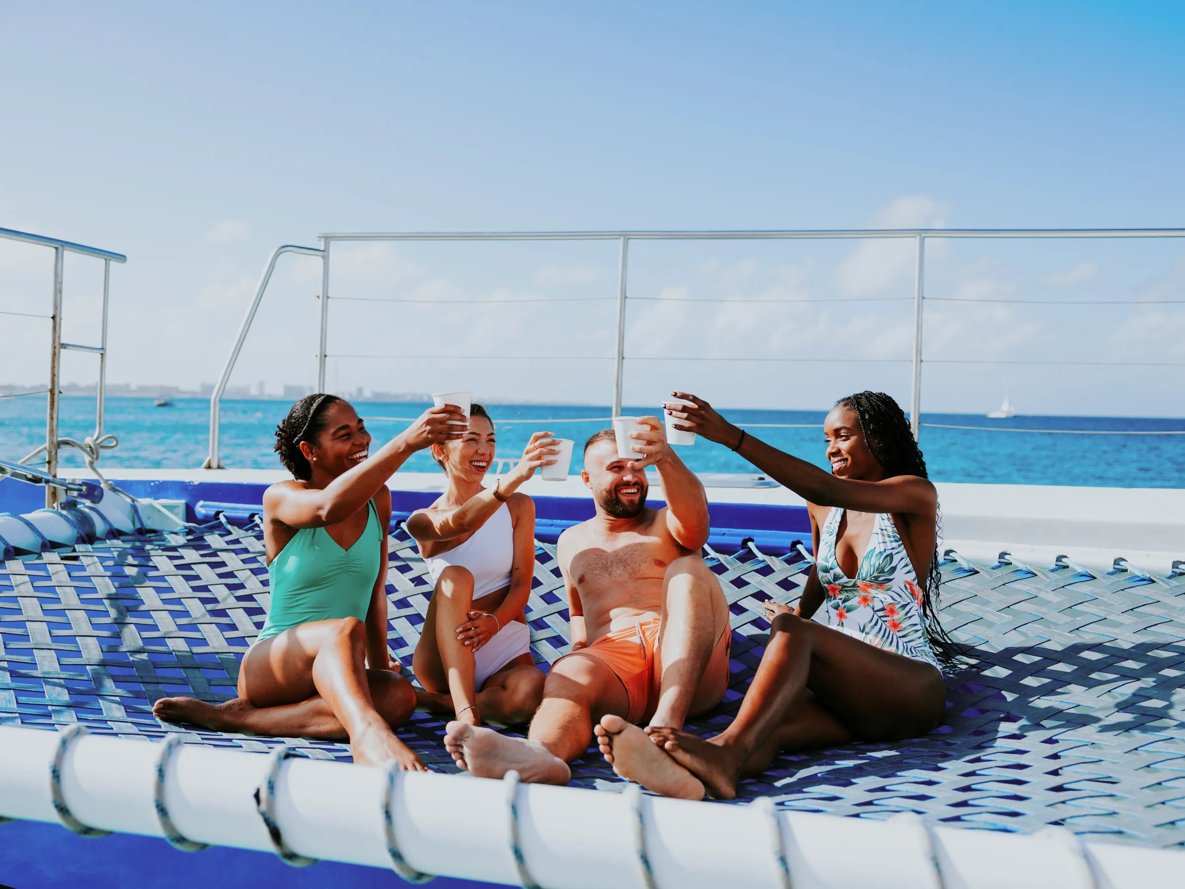 Group of four people in swimsuits toasting drinks on a catamaran net over the ocean.