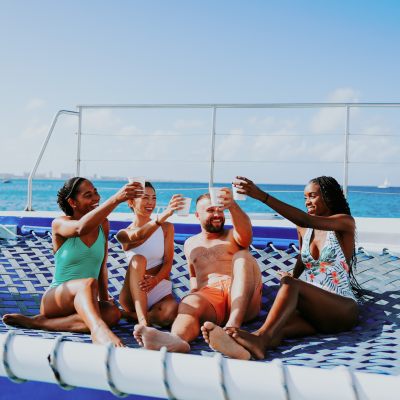 Group of four people in swimsuits toasting drinks on a catamaran net over the ocean.