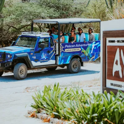 Tour group in an open-air blue jeep, 'Pelican Adventures', near a wooden sign marked 'AYO'.
