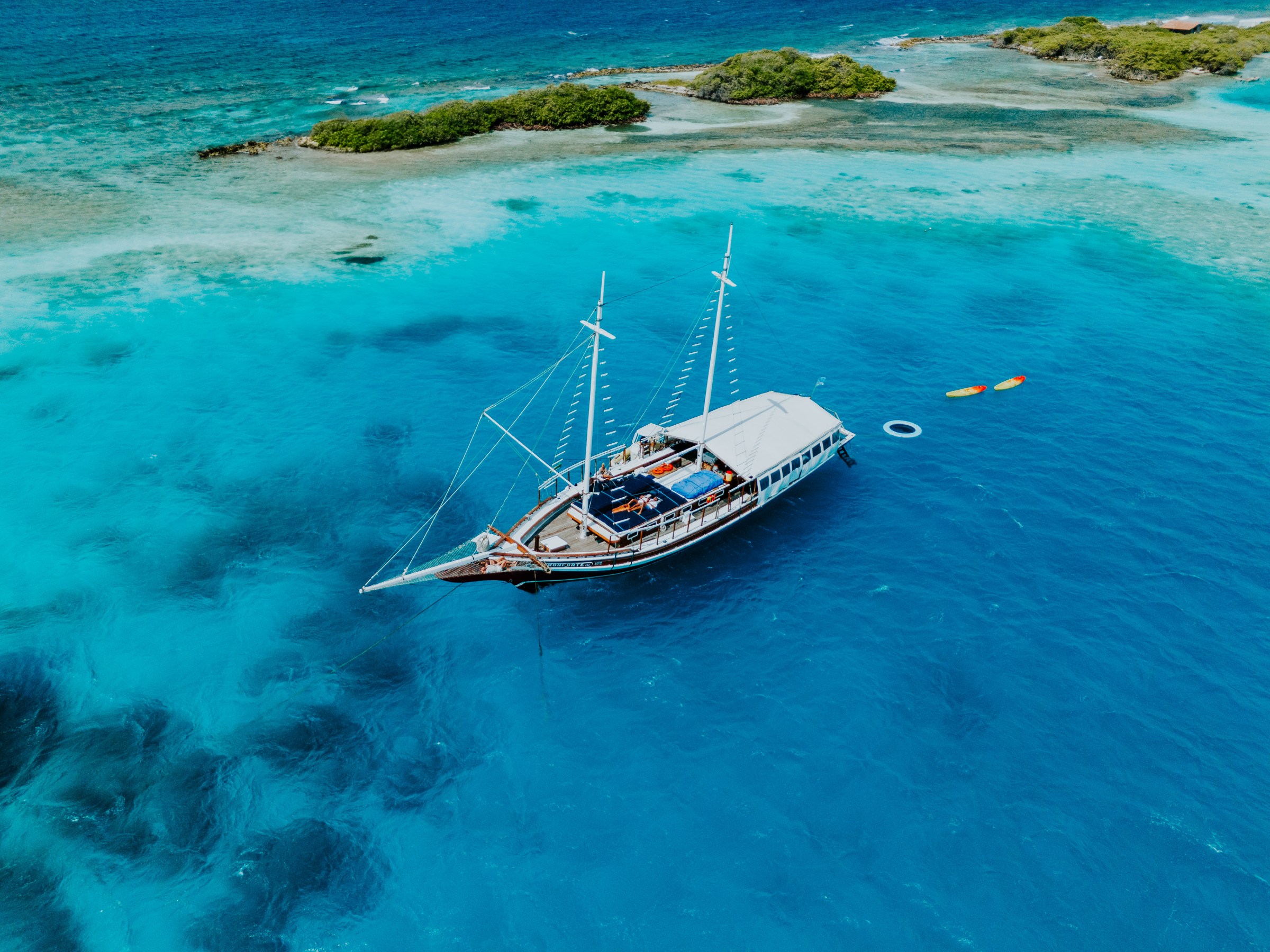 A sailboat anchored near a tropical island with clear blue water and small kayaks nearby.