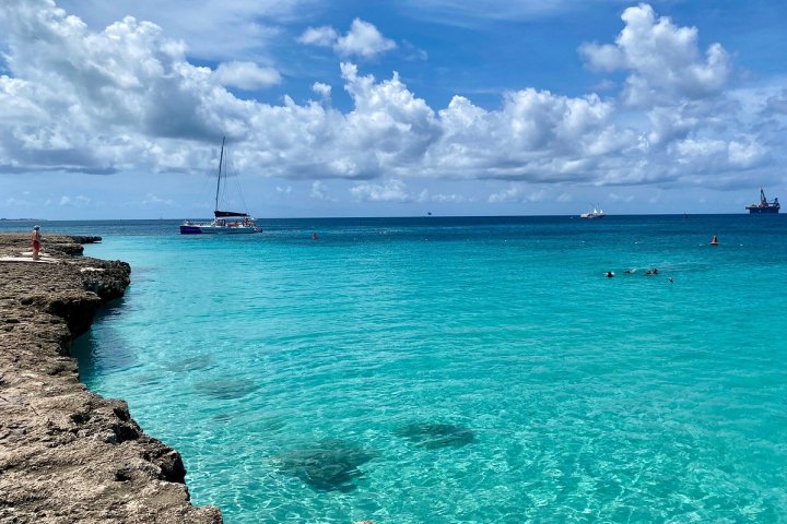 Turquoise sea with boats and swimmers, rocky shore in foreground, cloudy sky above.