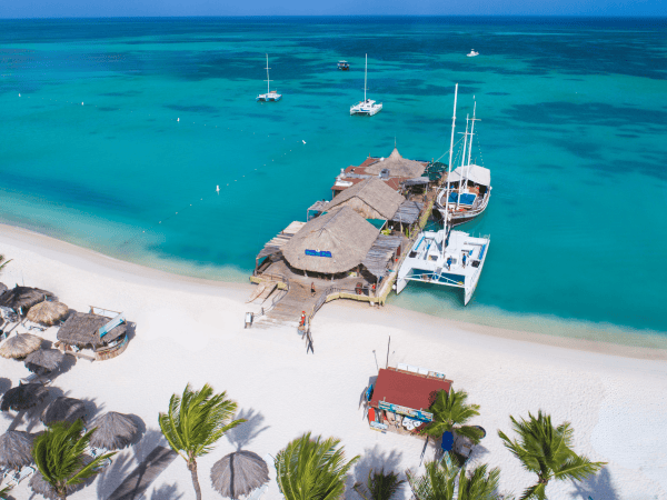 Tropical beach with huts, palm trees, turquoise water, and anchored sailboats.